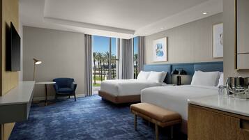 Habitación de hotel con dos camas, un escritorio, una silla, un banco, un televisor y vistas a palmeras.