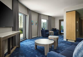 Habitación de hotel moderna con un ventanal grande, televisor de pantalla plana, sofá, dos sillones, una mesita redonda y un mueble de madera.
