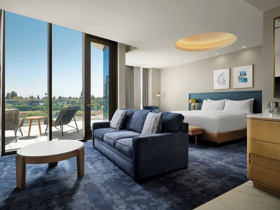 A hotel room with a large window, a sofa, a bed, a desk, and a view of the outdoors.