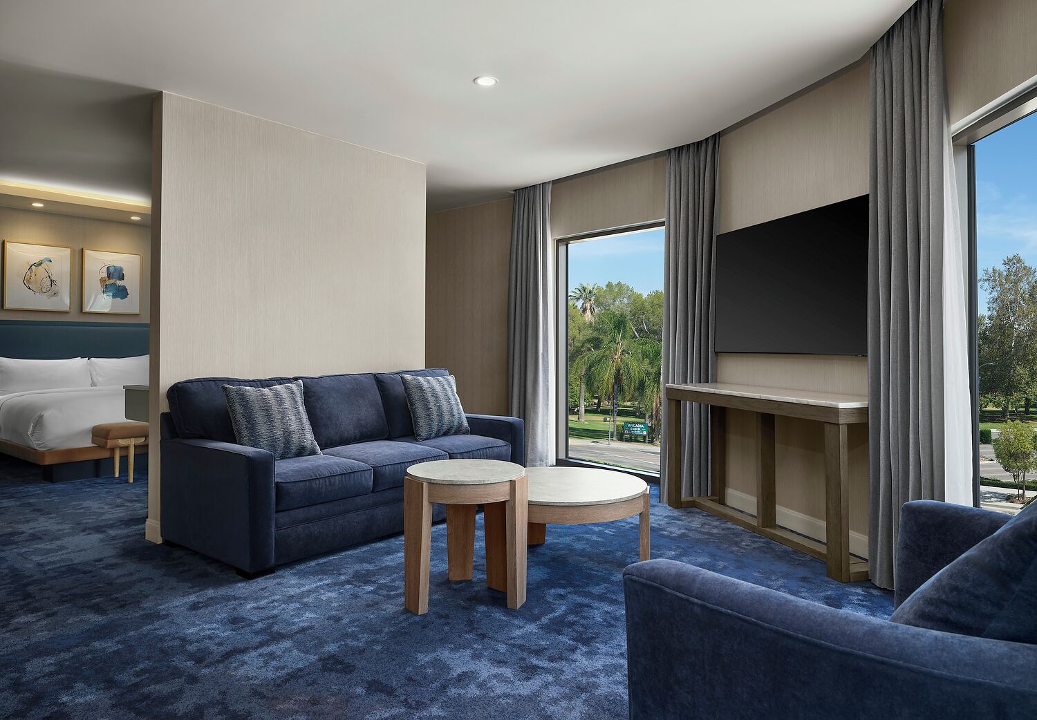 A modern hotel room with a large bed, a dark blue sofa, a round wooden table, and a TV mounted on the wall.