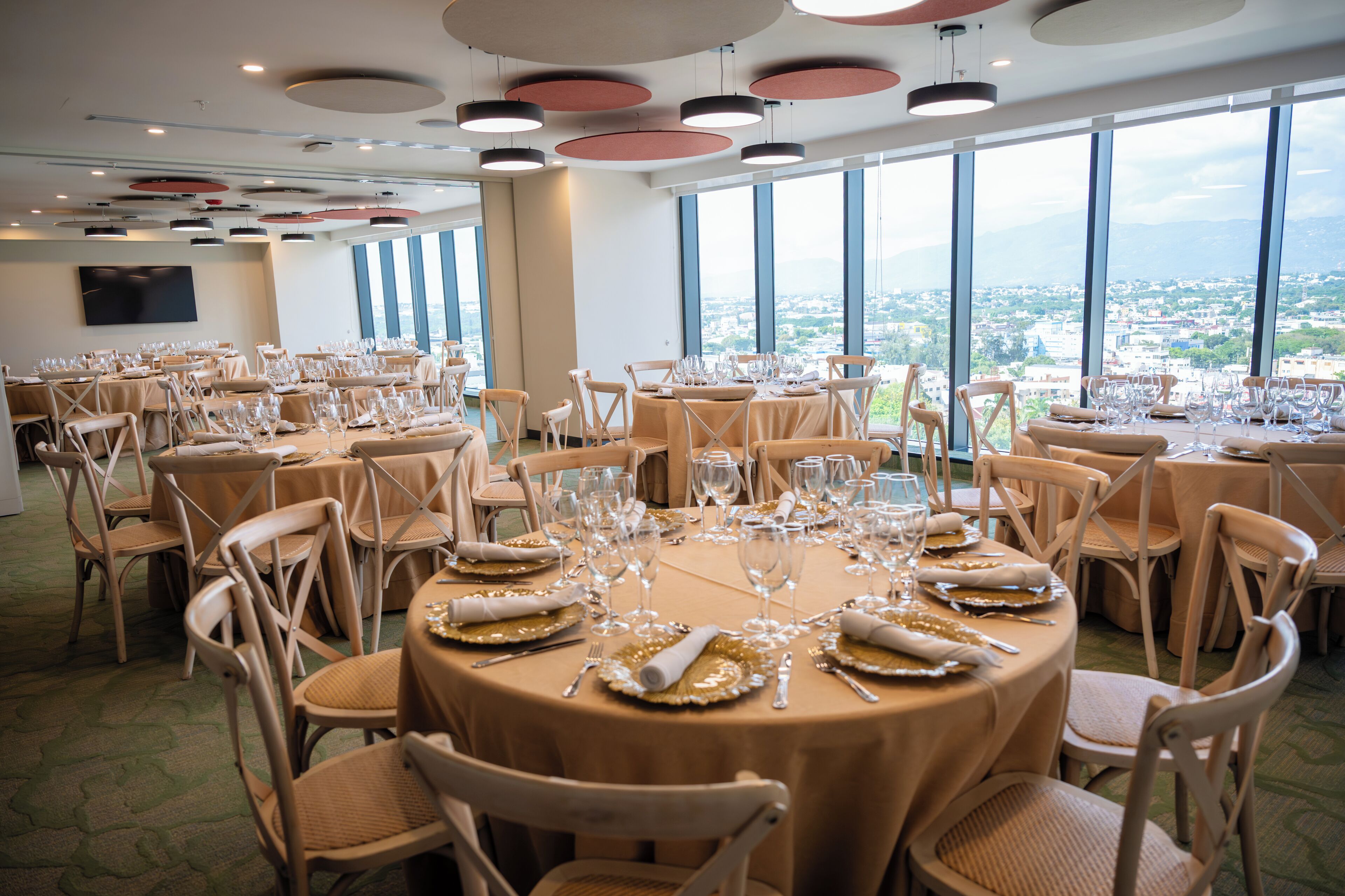 Sala de conferências com mesas redondas preparadas para um evento formal, grandes janelas com vista para a cidade e uma TV de tela plana na parede.
