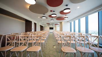 Un salón de conferencias con filas de sillas de madera y piso alfombrado, ventanas amplias en un lateral y luces circulares en el techo.