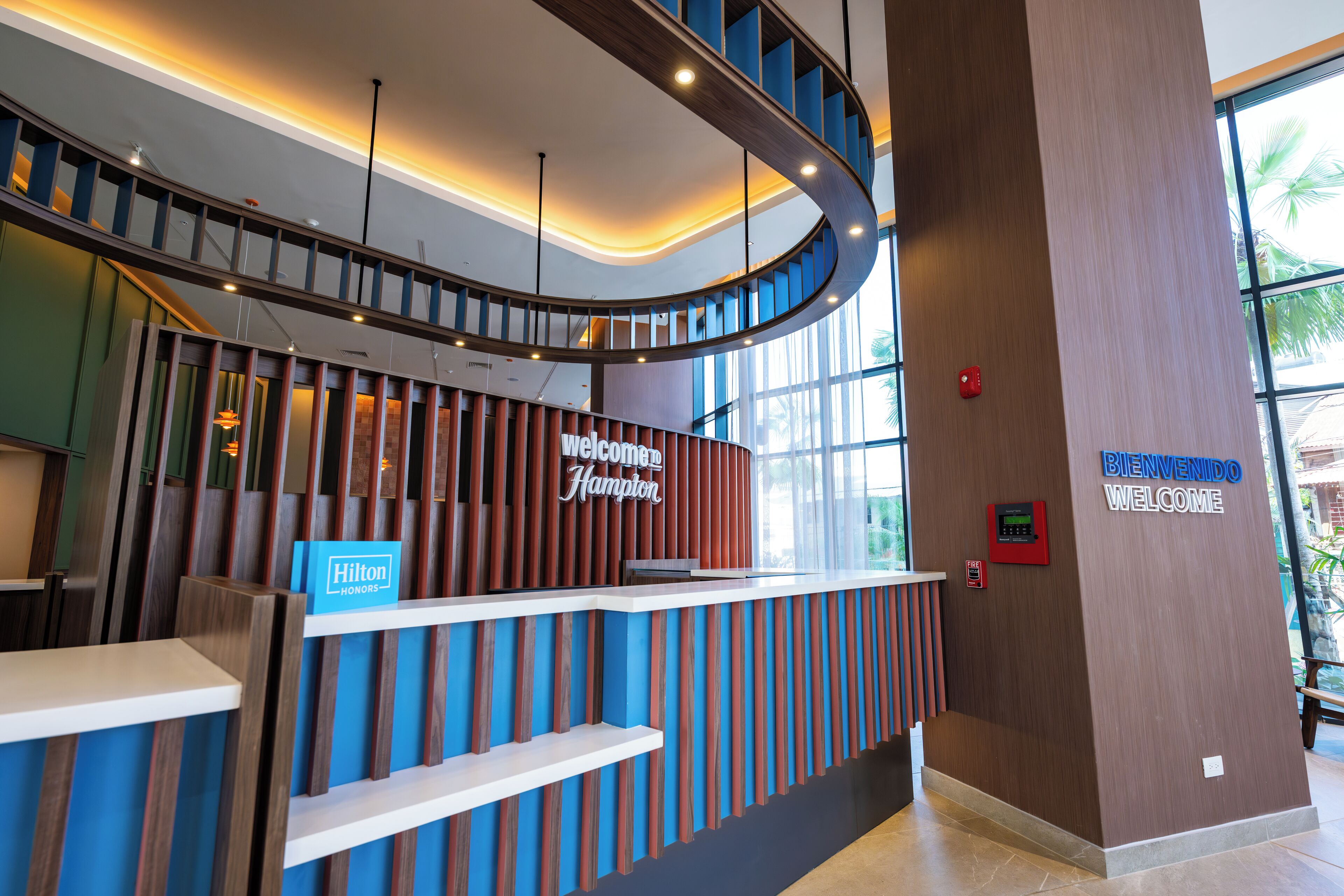 A modern hotel reception desk with a curved staircase, a 'Welcome to Hampton' sign, and a 'Hilton Honors' sign.