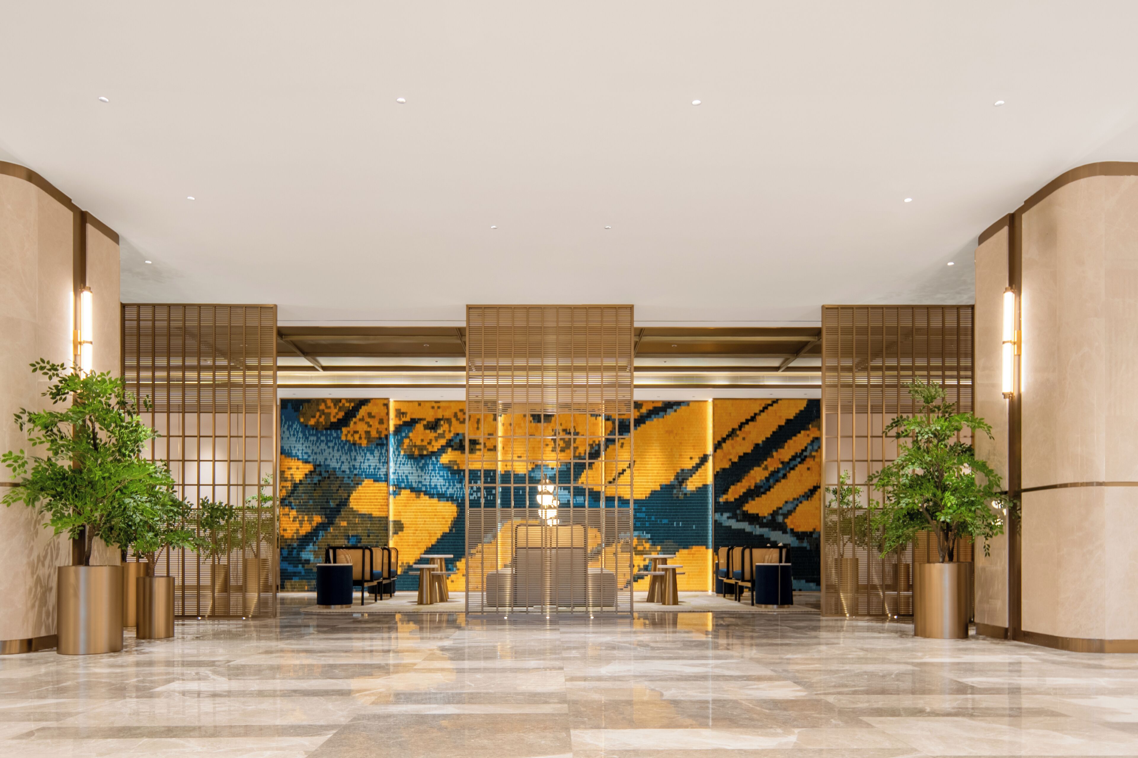 A spacious lobby with a modern design, featuring a large mosaic wall, seating areas, and potted plants.