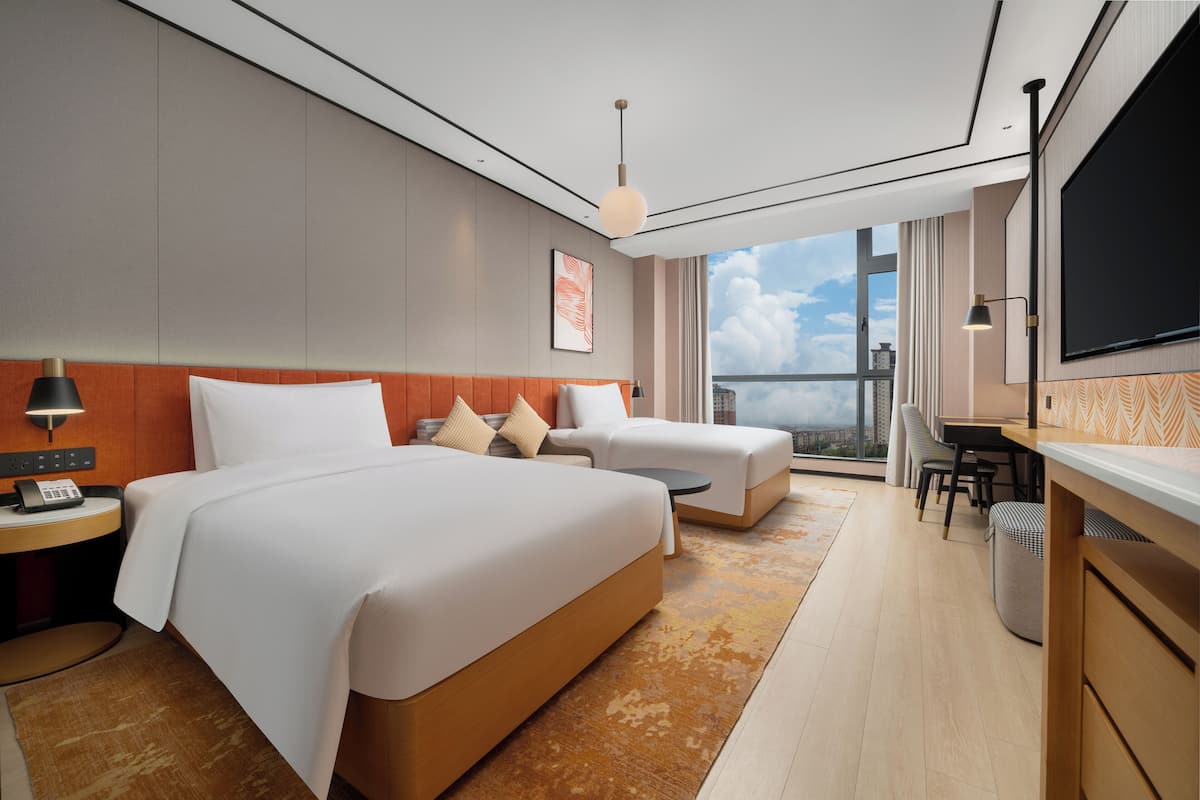 Habitación de hotel moderna con dos camas, un escritorio, un televisor y un ventanal con vistas a la ciudad.