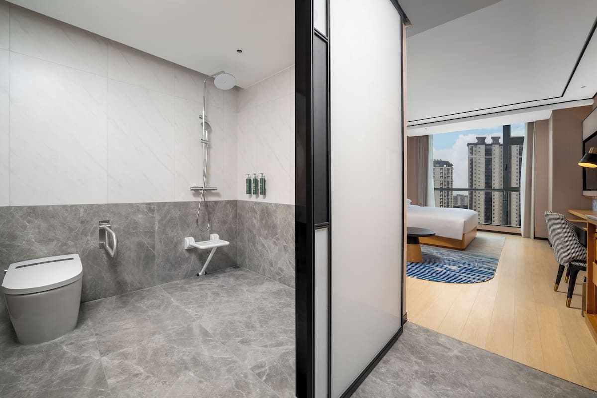 Un baño moderno con ducha, inodoro y lavamanos. En la habitación contigua hay una cama y un escritorio con vistas a edificios altos.