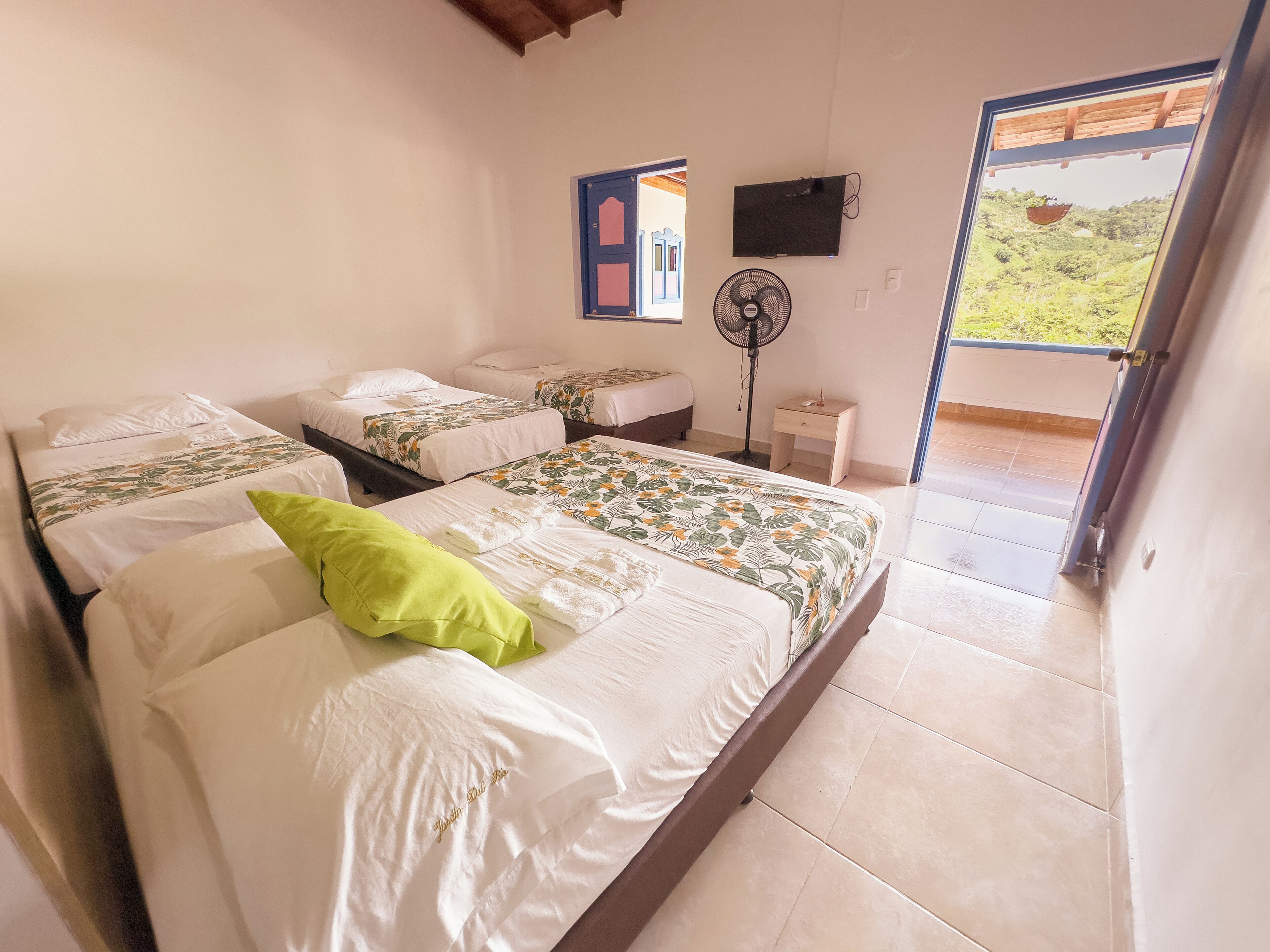 Habitación de hotel con dos camas, televisión, ventilador y balcón con vista a zonas verdes.