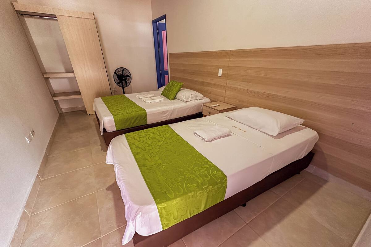 A hotel room with two beds, each with white linens and green accents, a wooden headboard, a small bedside table, and a wall-mounted fan.