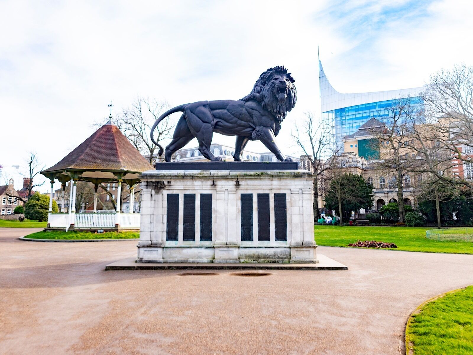 A lion statue in a park with a gazebo and a modern building in the background.