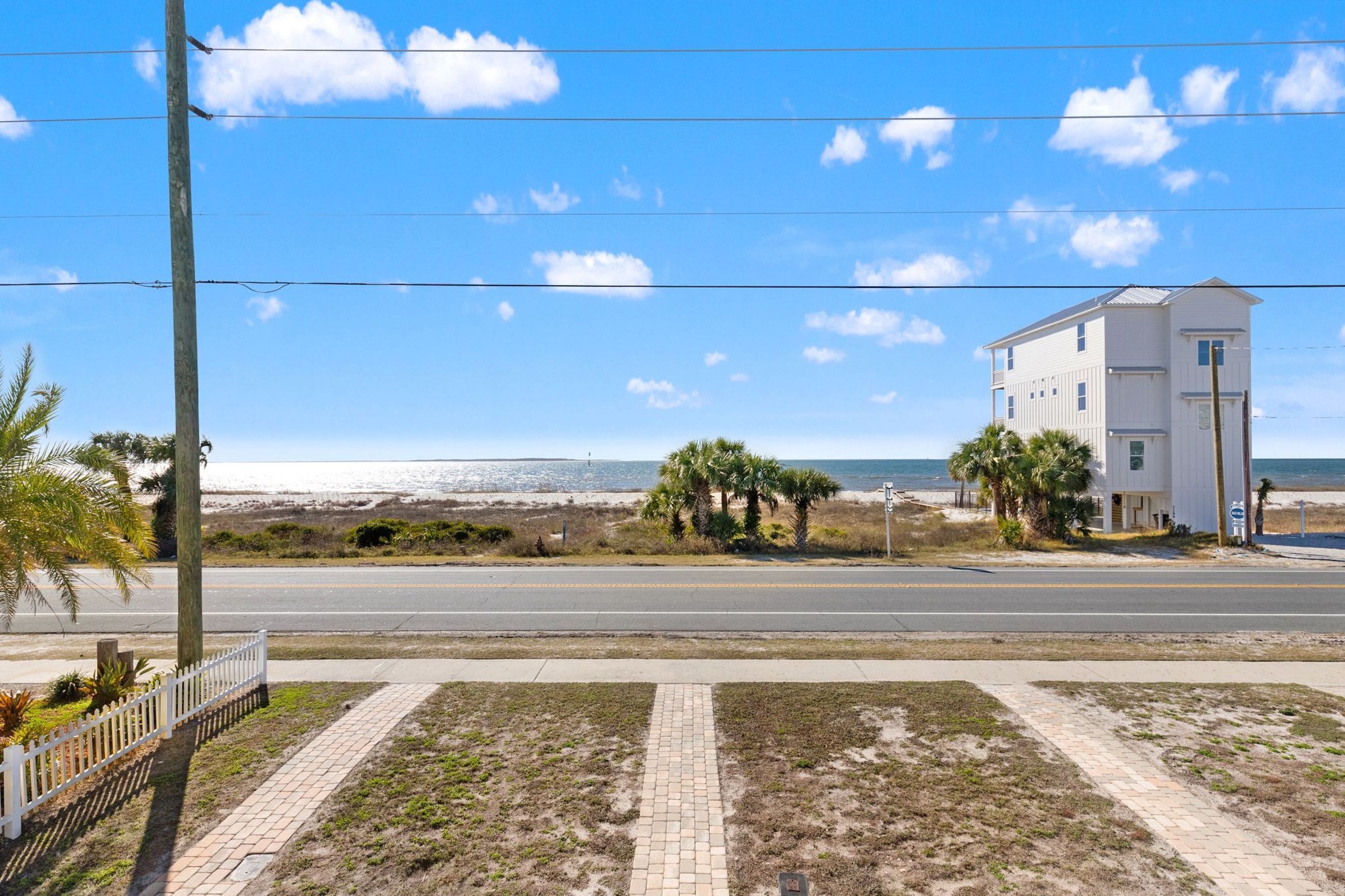 Una carretera costera con un edificio blanco, palmeras y una playa al fondo.