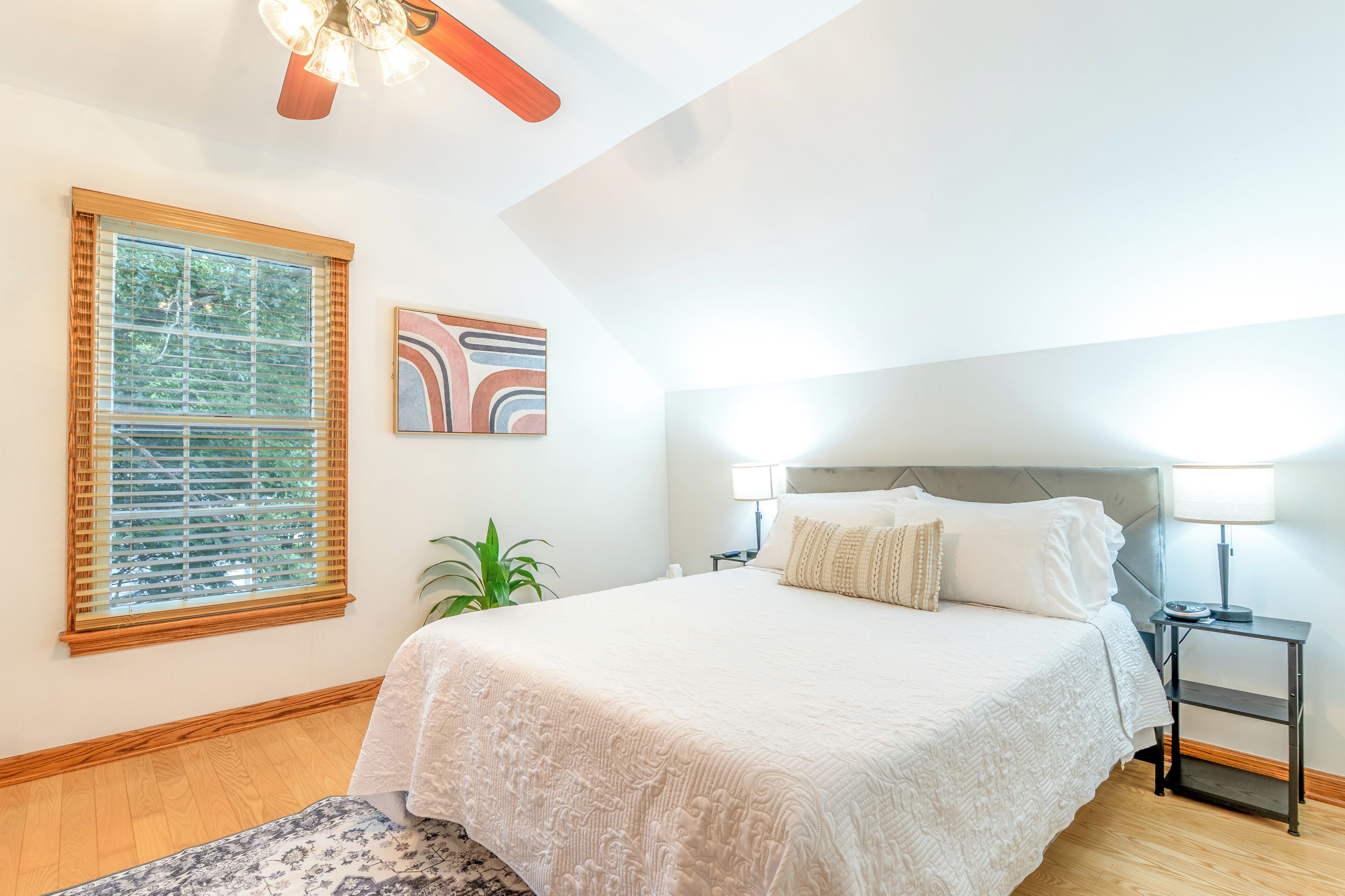 A bedroom with a large bed, a nightstand, a lamp, a window with blinds, and a ceiling fan.