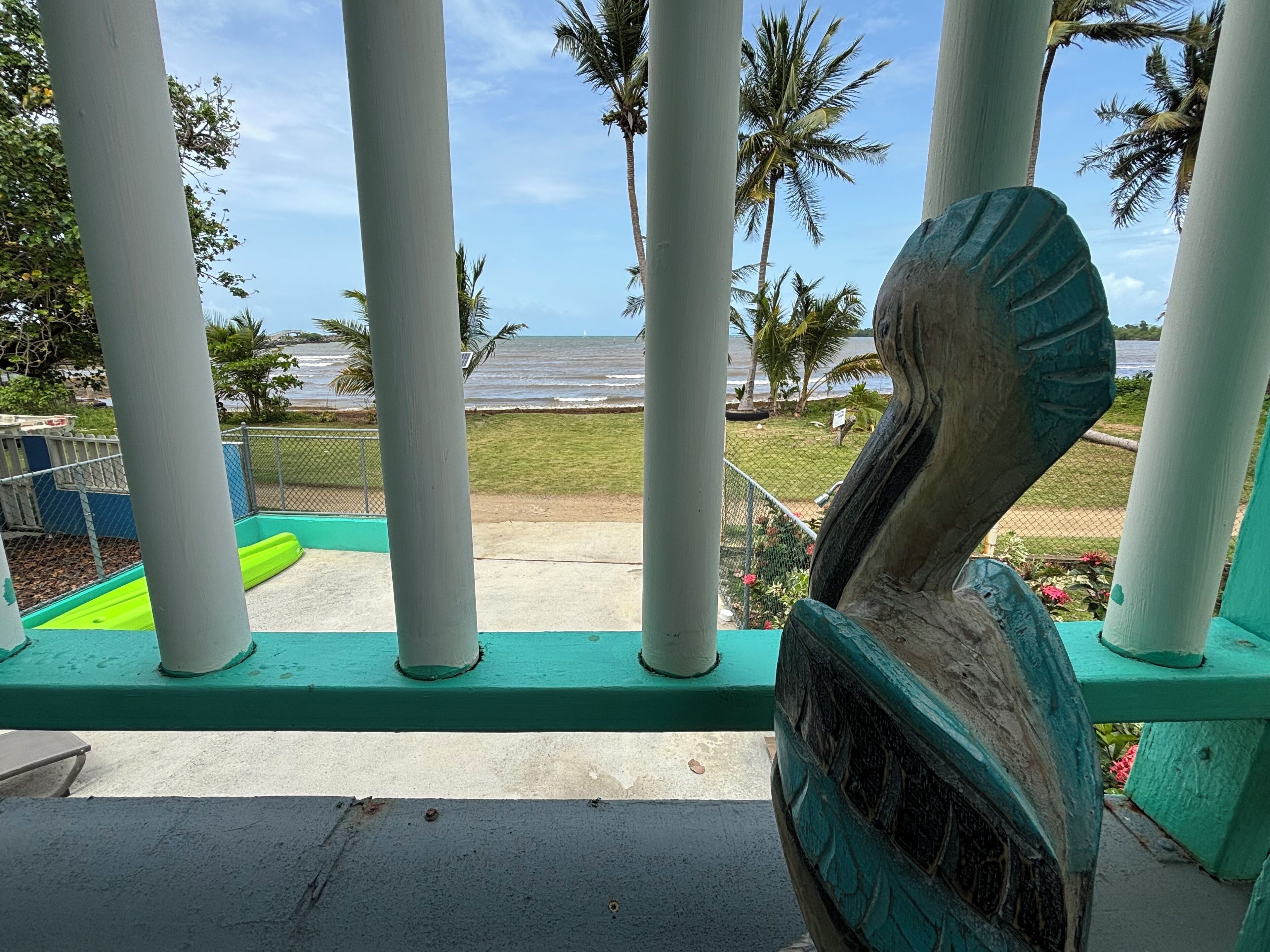 Une vue depuis un balcon doté d’une balustrade, donnant sur une plage bordée de palmiers et un ciel dégagé.