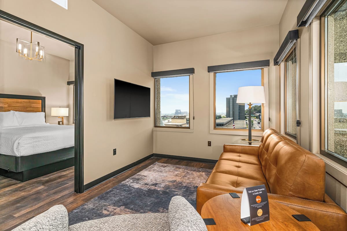 A hotel room with a bed, a sofa, a TV, and large windows with a city view.