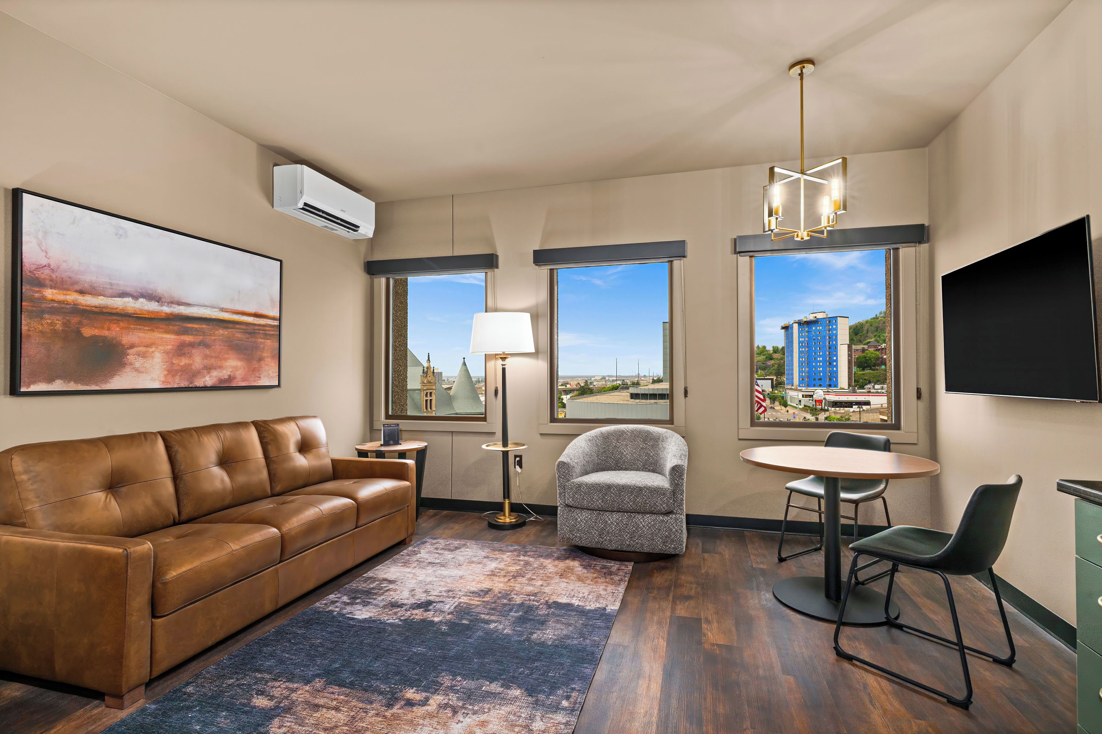 A modern hotel room with a leather sofa, a round dining table, a chair, a floor lamp, and a large window with a city view.