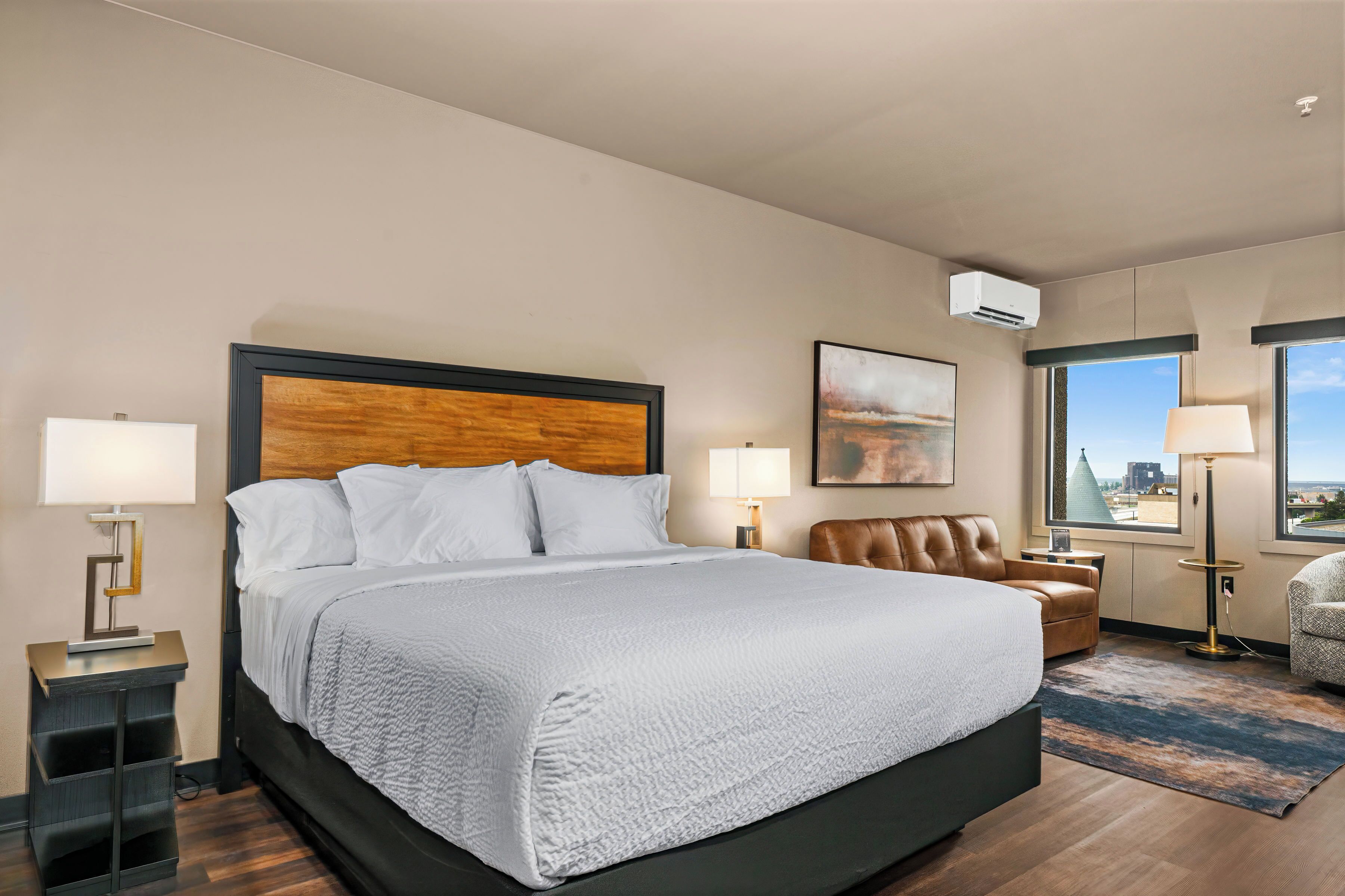 A hotel room with a large bed, a desk with a lamp, a chair, and a view of the city through the window.