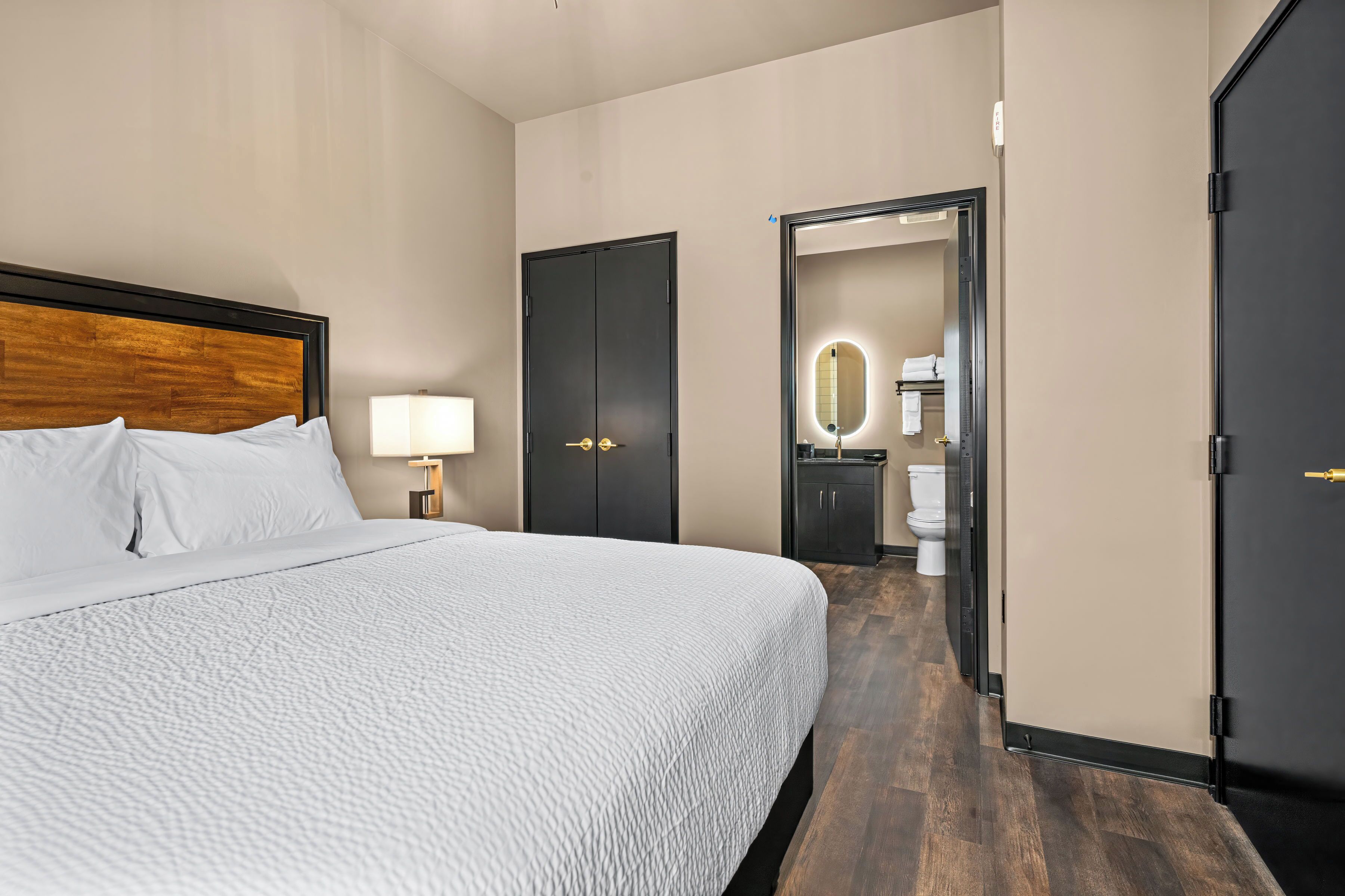 A hotel room with a large bed, a nightstand with a lamp, a wardrobe, and a bathroom visible through an open door.