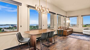 A modern living room with a wooden dining table, two chairs, a brown leather sofa, and a gray armchair. Large windows offer a view of a body of water and buildings.