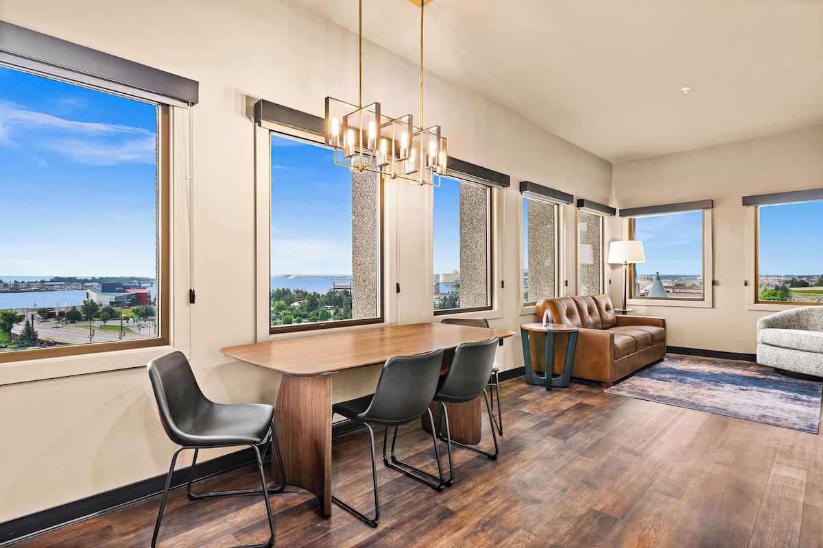 A modern living room with a wooden dining table, two chairs, a brown leather sofa, and a gray armchair. Large windows offer a view of a body of water and buildings.