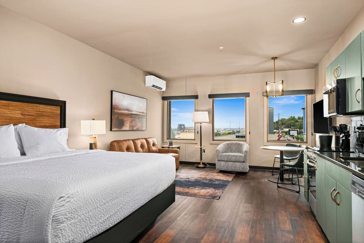 A hotel room with a large bed, a desk with a chair, a kitchenette, and a view of the city through large windows.
