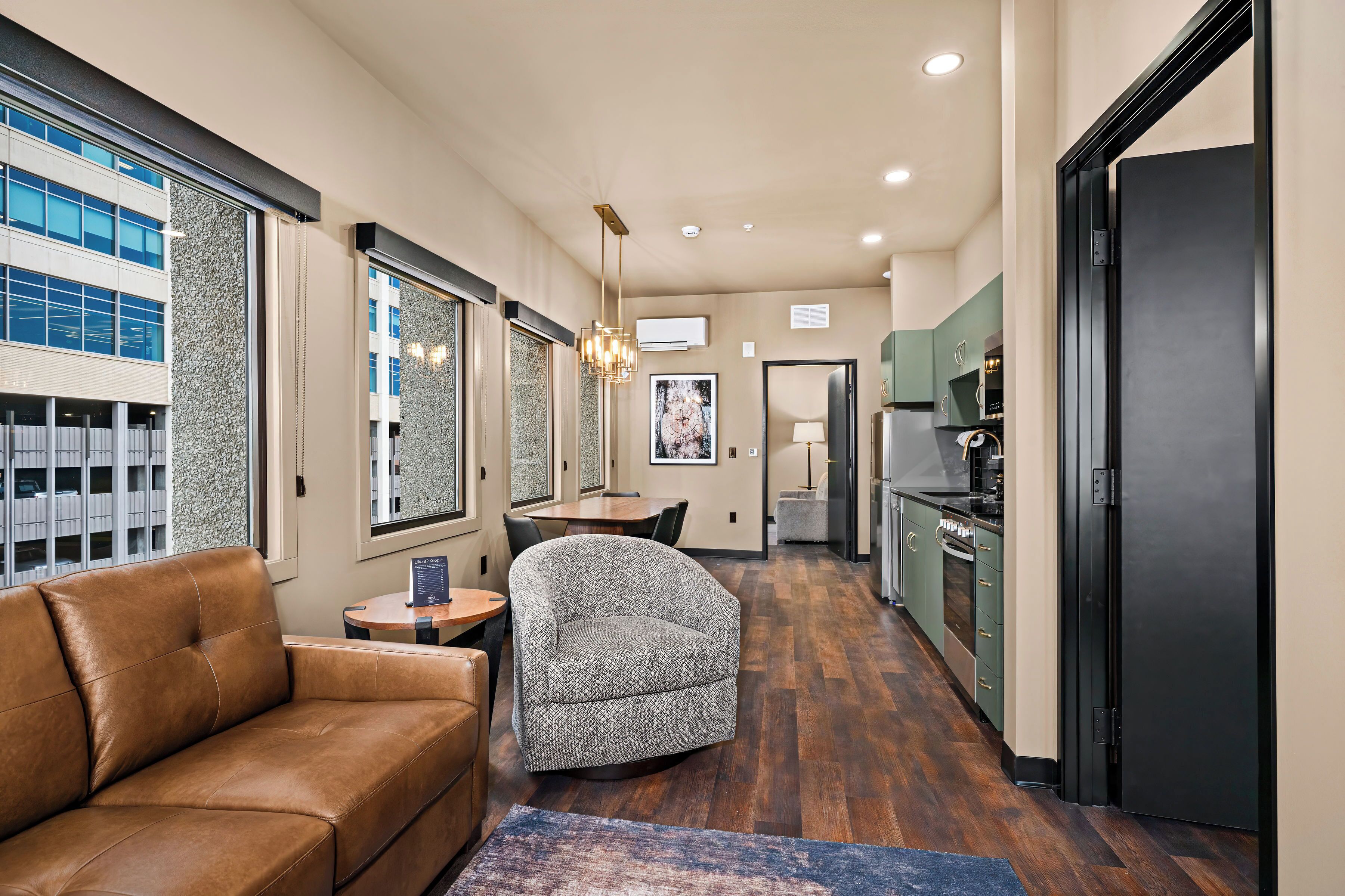 A modern kitchen with a black refrigerator, a dining area with a table and chairs, and a living area with a brown leather sofa and a patterned armchair.