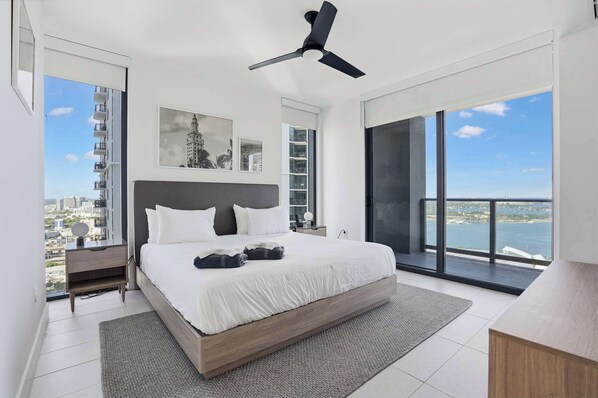 Une chambre moderne avec un grand lit, un ventilateur de plafond, une vue sur la ville et un balcon donnant sur la mer.