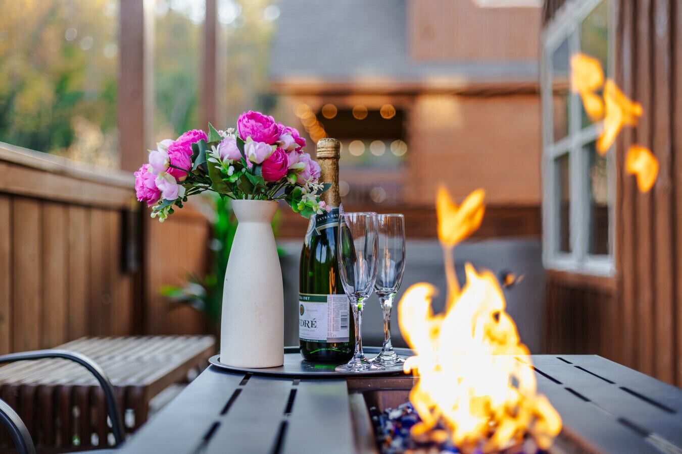 A table with a bottle of wine, two glasses, and a vase of flowers on a wooden deck.