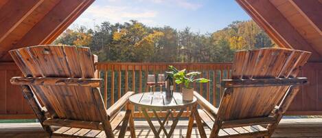 A wooden deck with two Adirondack chairs and a small table with a potted plant and wine glasses.