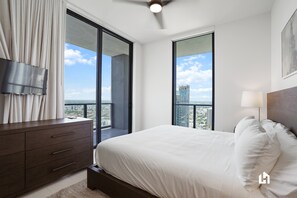 A modern bedroom with a large bed, a TV mounted on the wall, and a balcony with a city view.