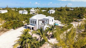 Una casa blanca de dos pisos con un porche cubierto y una piscina, rodeada de vegetación tropical y otras viviendas.