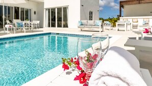Un área junto a la piscina con un vaso de agua, flores, muebles blancos y un edificio moderno al fondo.