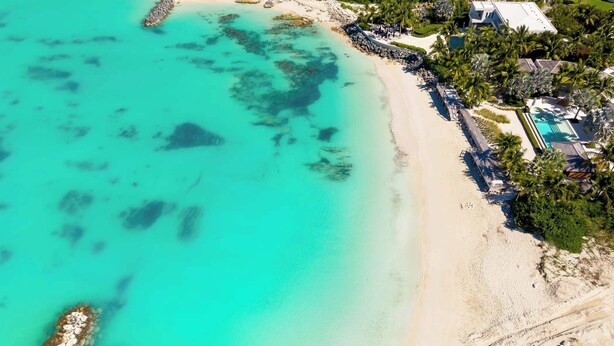 Vista aérea de un complejo turístico en la playa, con aguas turquesas cristalinas, una orilla de arena y una zona de piscina rodeada de palmeras y edificios del complejo.