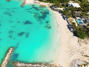 An aerial view of a beach resort with clear turquoise waters, a sandy shoreline, and a pool area surrounded by palm trees and resort buildings.