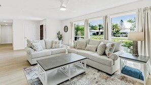 Ein geräumiges Wohnzimmer mit einer großen, im L-förmigen Winkel stehenden Couch, einem Couchtisch und einem Beistelltisch mit einer Lampe. Große Fenster bieten Blick auf einen Garten und ein Haus.