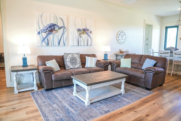 A living room with a brown leather sectional sofa, a wooden coffee table, and a blue rug. There are two side tables with lamps, a wall clock, and abstract paintings.