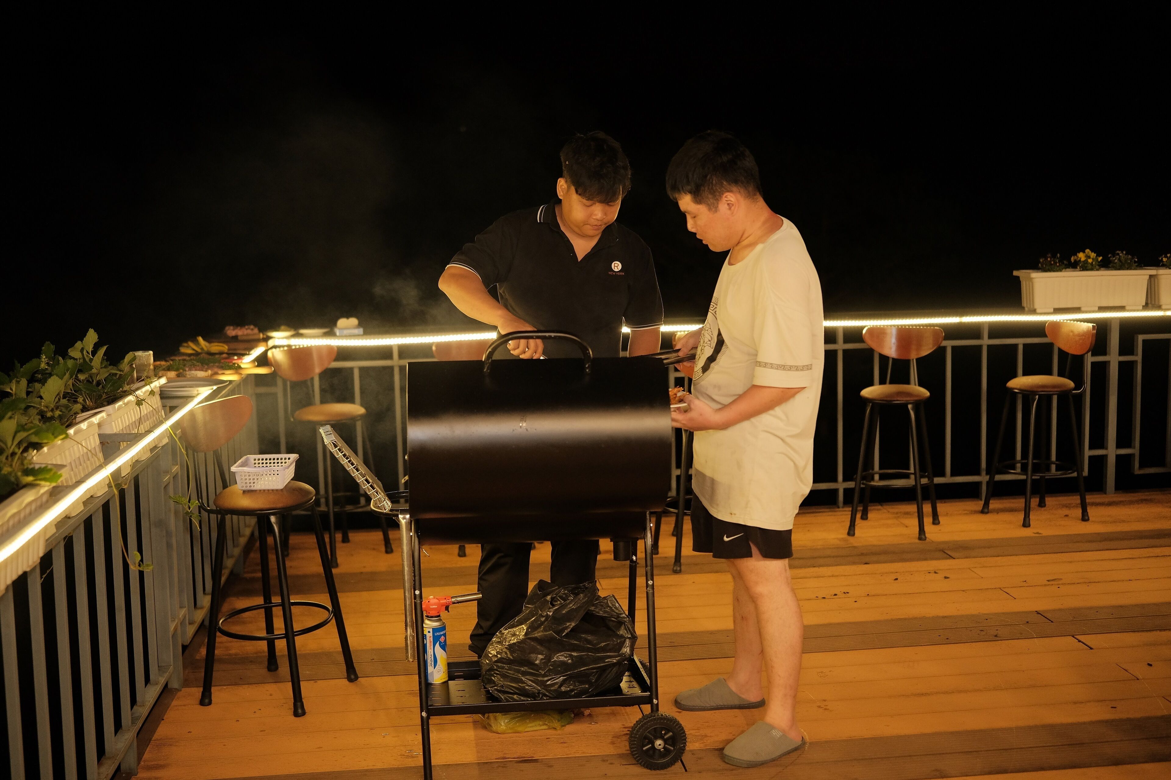 Two people grilling outdoors at night with a bar area and stools in the background.