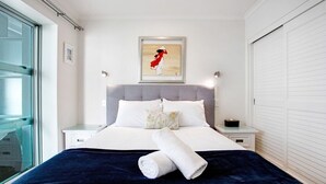 A neatly made bed with a dark blue blanket and a rolled white towel, a framed picture on the wall, and a white headboard. - Space Style and Comfort (Auckland)