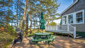 En træterrasse med et picnicbord og en parasol, omgivet af høje træer og et hus med et hvidt gelænder.