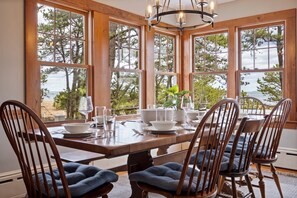 A dining area with a wooden table set for four, large windows with a view of trees and water, and a chandelier above. - Beachfront • 30 Steps to the Beach • Sunrise Views (Saco)