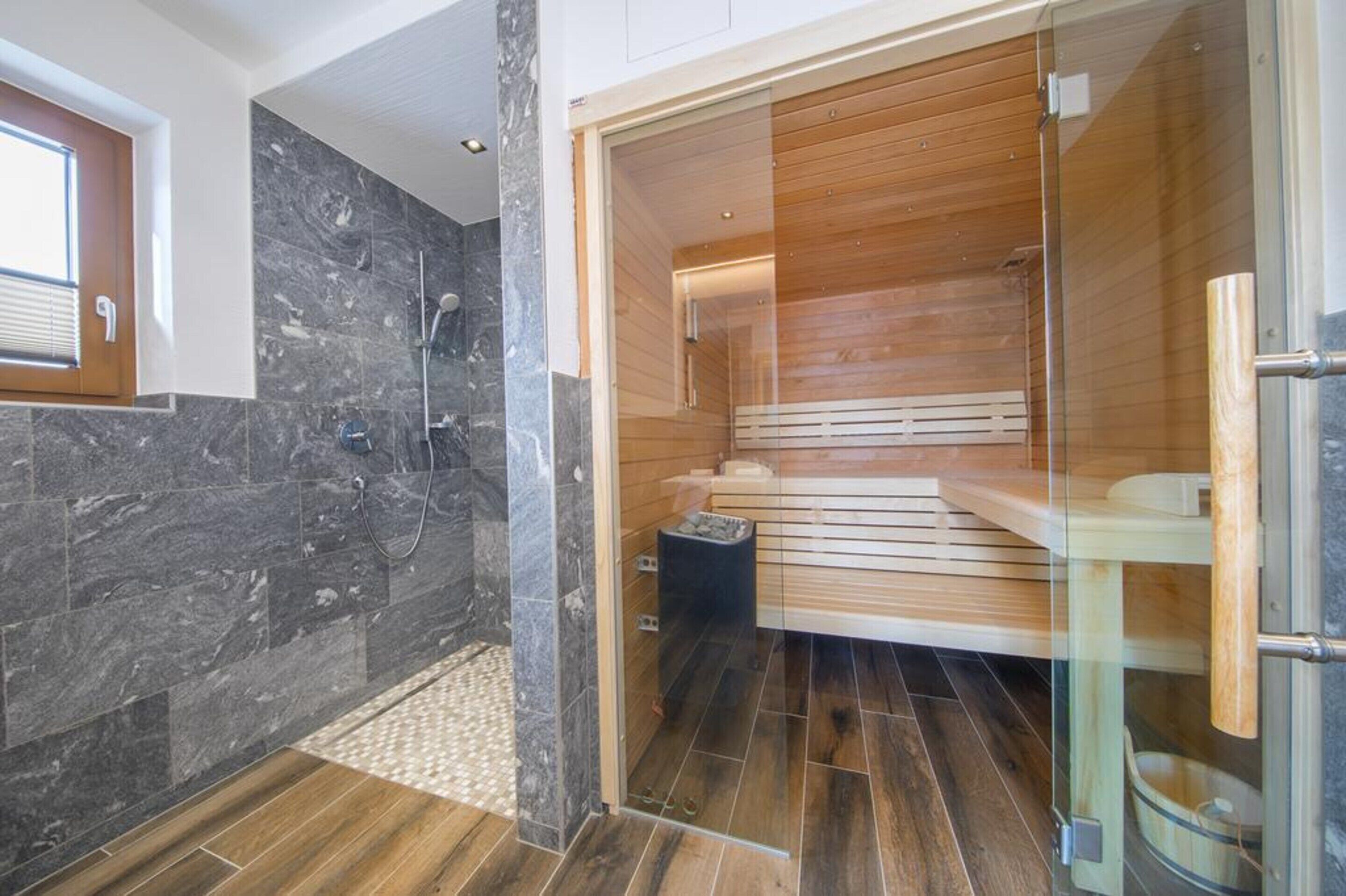A modern bathroom with a glass-enclosed sauna, a shower area with marble tiles, and a wooden floor.
