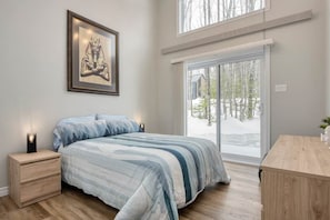 A bedroom with a bed, a nightstand, a wooden desk, a window with a view of trees, and a framed picture on the wall.