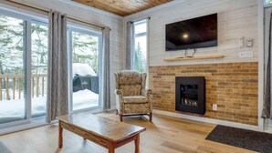 A living room with a fireplace, a television mounted above it, and a large window with curtains. - Hôtel à la maison - Le Cocon, ski et golf (Adstock)