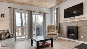 A living room with a sofa, a coffee table, a fireplace, a TV mounted above it, and a chair. - Hôtel à la maison - La Cachette, golf et ski! (Adstock)
