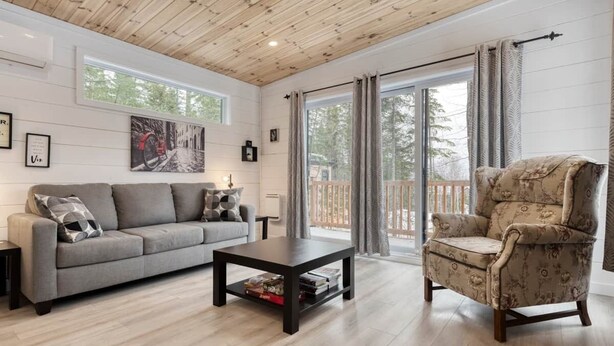 A living room with a sofa, armchair, coffee table, and a large window with curtains. - Hôtel à la maison - La Cachette, golf et ski! (Adstock)