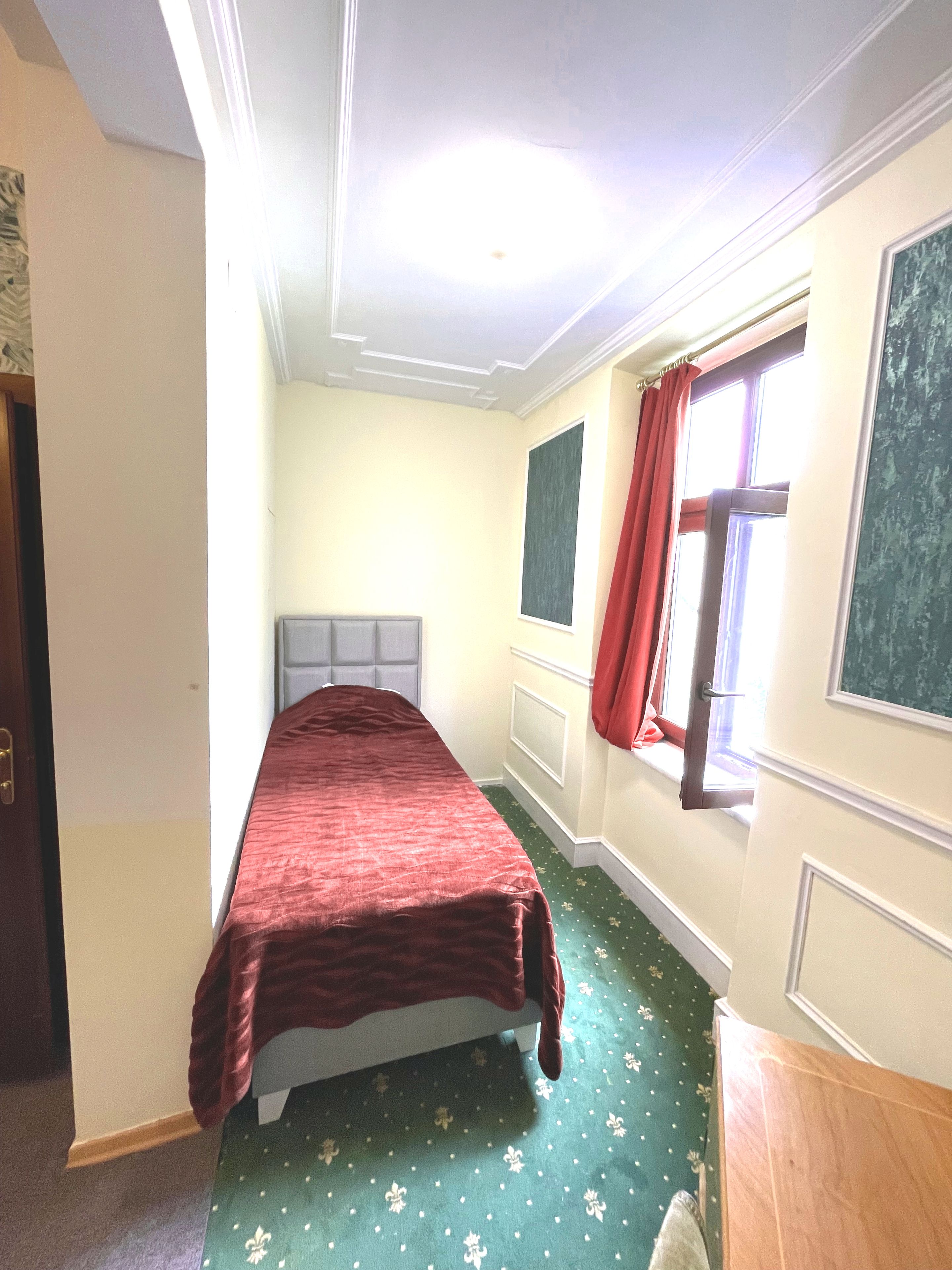 Ein Zimmer mit einem Bett, einem Fenster mit roten Vorhängen und einem grünen Teppich mit Muster.