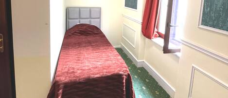 Ein Zimmer mit einem Bett, einem Fenster mit roten Vorhängen und einem grünen Teppich mit Muster.