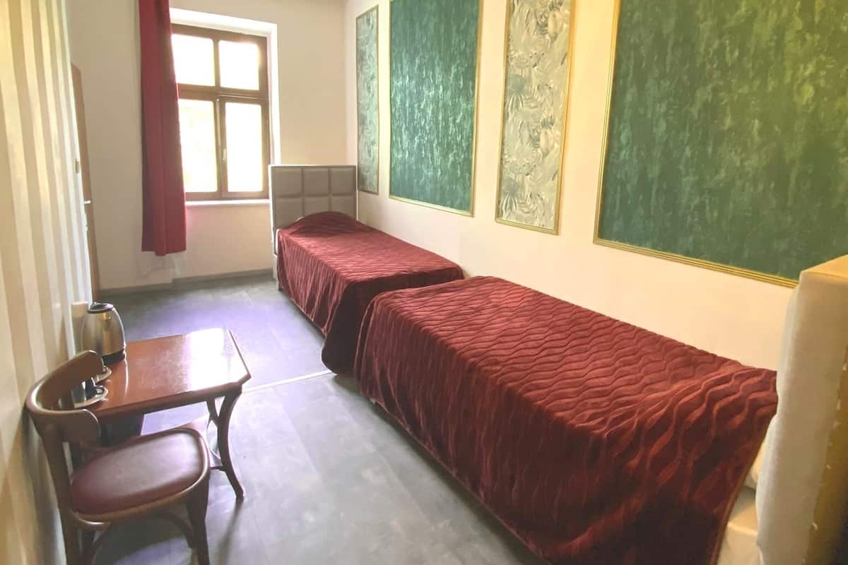 A dormitory room with two single beds, a wooden table with a chair, a radiator, and a window with a red curtain.