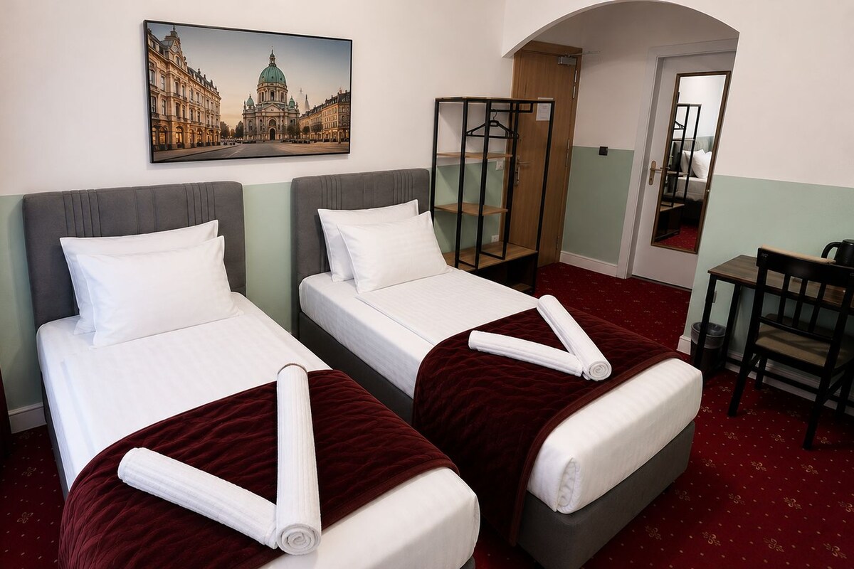 A hotel room with two beds, a desk, a chair, and a painting of a building.