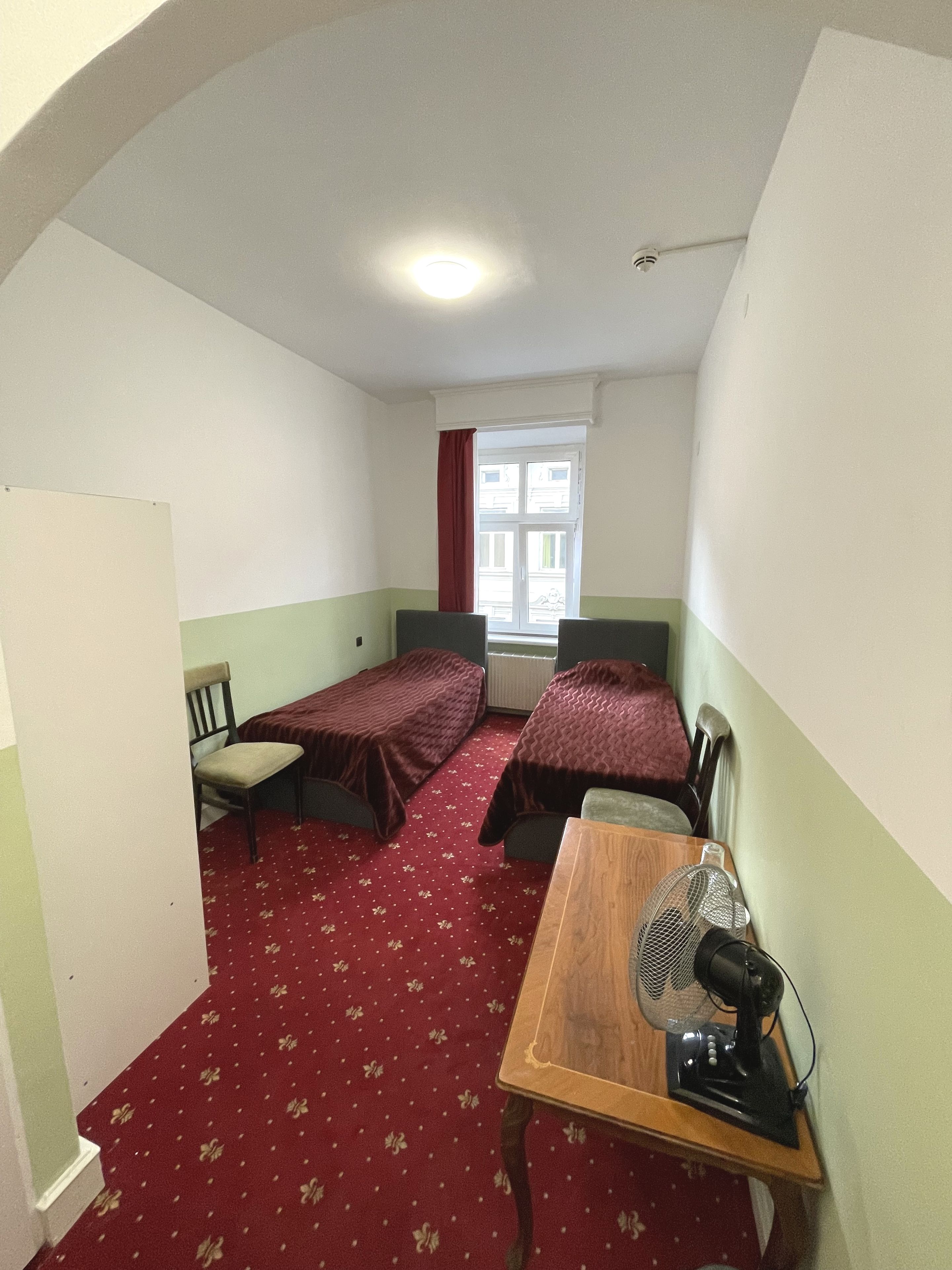 A dormitory room with two single beds, a wooden table with a fan, a chair, and a window with a red curtain.