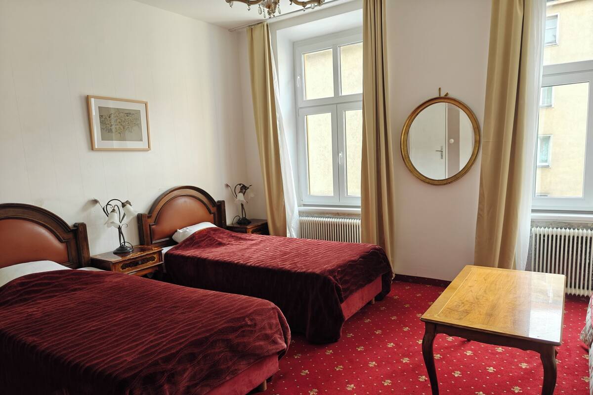 A hotel room with two beds, a wooden table, a round mirror, and a window with curtains.