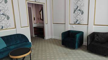 Ein Zimmer mit einem türkisfarbenen Sofa, einem dunkelgrünen Sessel und einem schwarzen Sessel. Vor dem Sofa steht ein kleiner Holztisch. Die Wände sind mit floralem Wandpapier mit goldener Zierleiste versehen.